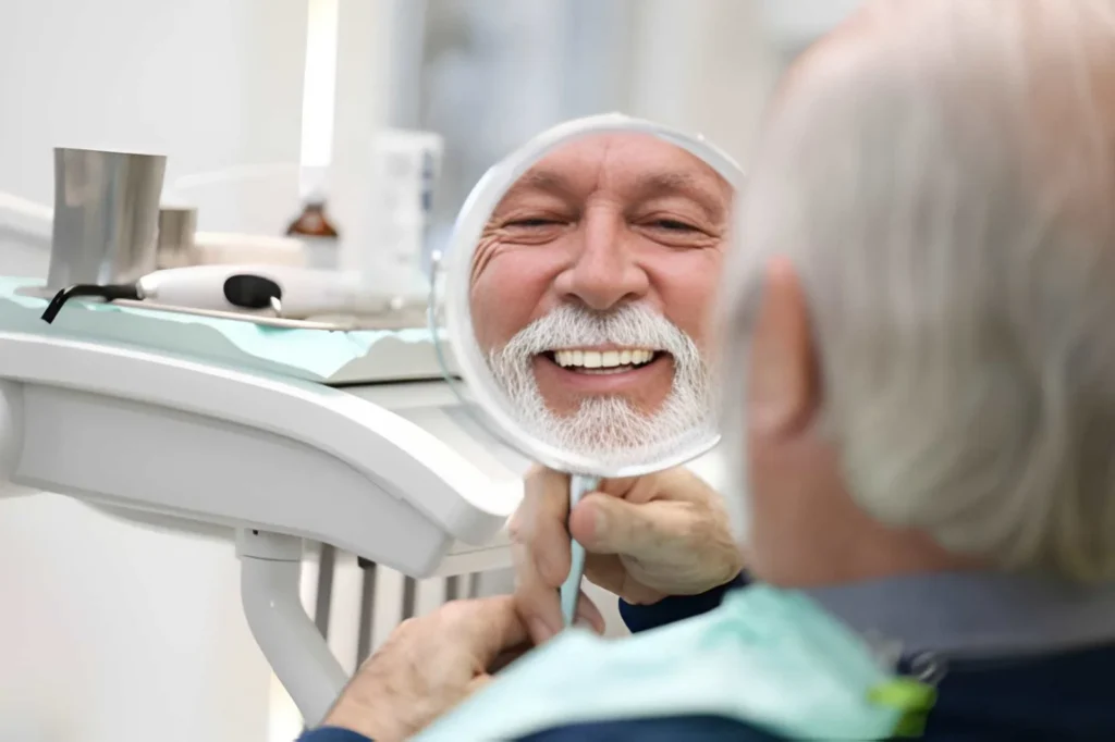 Older dental patient smiling after reviewing denture treatment results in the mirror